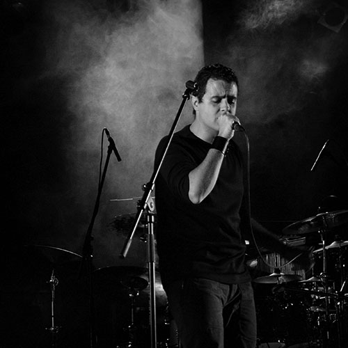 Tiago singing live. There's a microphone in his hand, a microphone stand in front of him and his eyes are closed. Behind him there's part of a drumkit and another microphone stand.